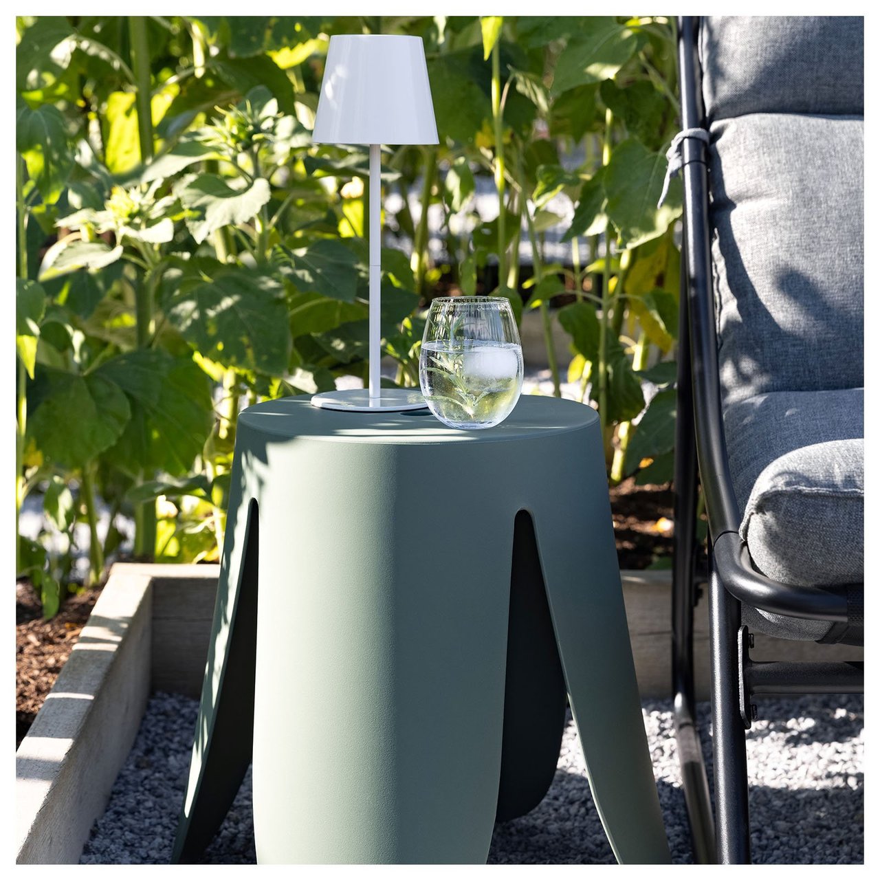 En grønn krakk står ved siden av en lenestol på en uteplass. På krakken står det en lampe og et glass.