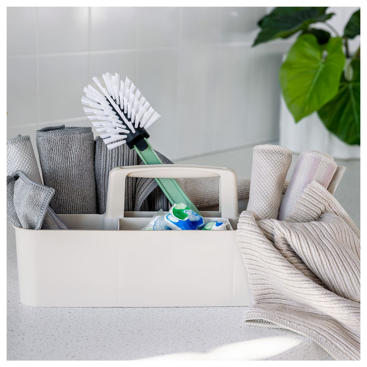 Nahaufnahme einer beigen Aufbewahrungsbox mit Griff auf einer weißen Spüle. Die Box enthält eine Spülbürste, Mikrofasertücher und Spültabs.