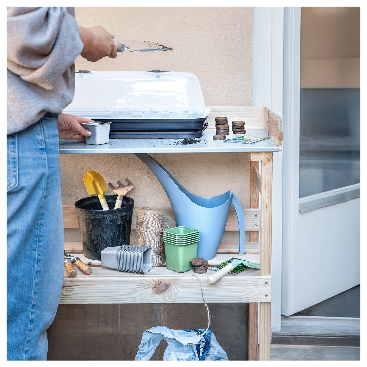 En person ved et plantebord med minidrivhus, vannkanne og dyrkingstilbehør.
