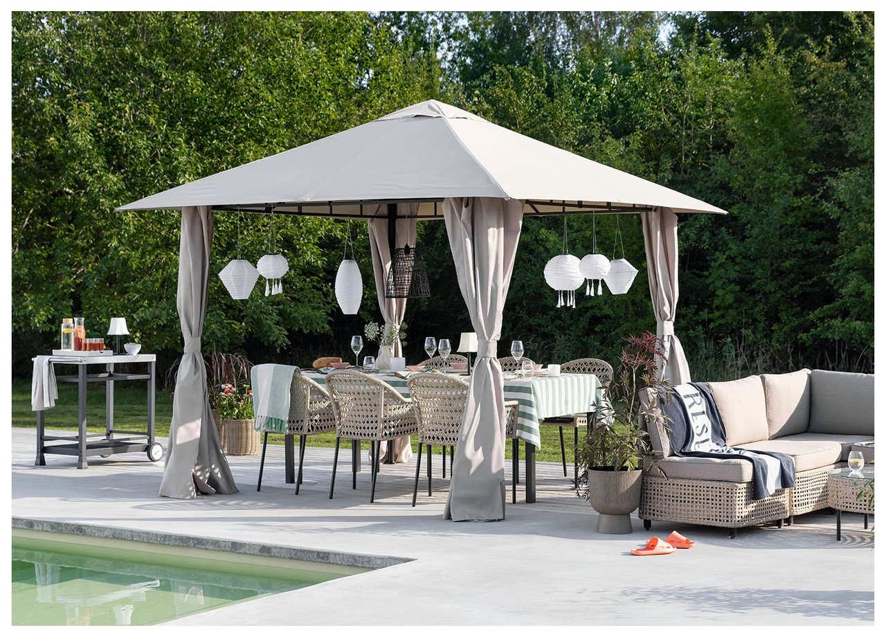 Ein großer Pavillon auf einem Rasen an einem See wirft Schatten über einen Loungebereich, der aus einem Sofa und Tisch aus Kunstrattan besteht.