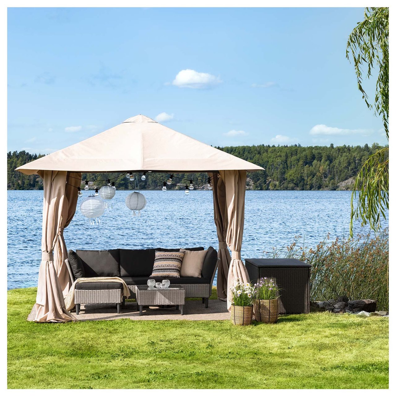 Ein großer Pavillon auf einem Rasen an einem See wirft Schatten über einen Loungebereich, der aus einem Sofa und Tisch aus Kunstrattan besteht.