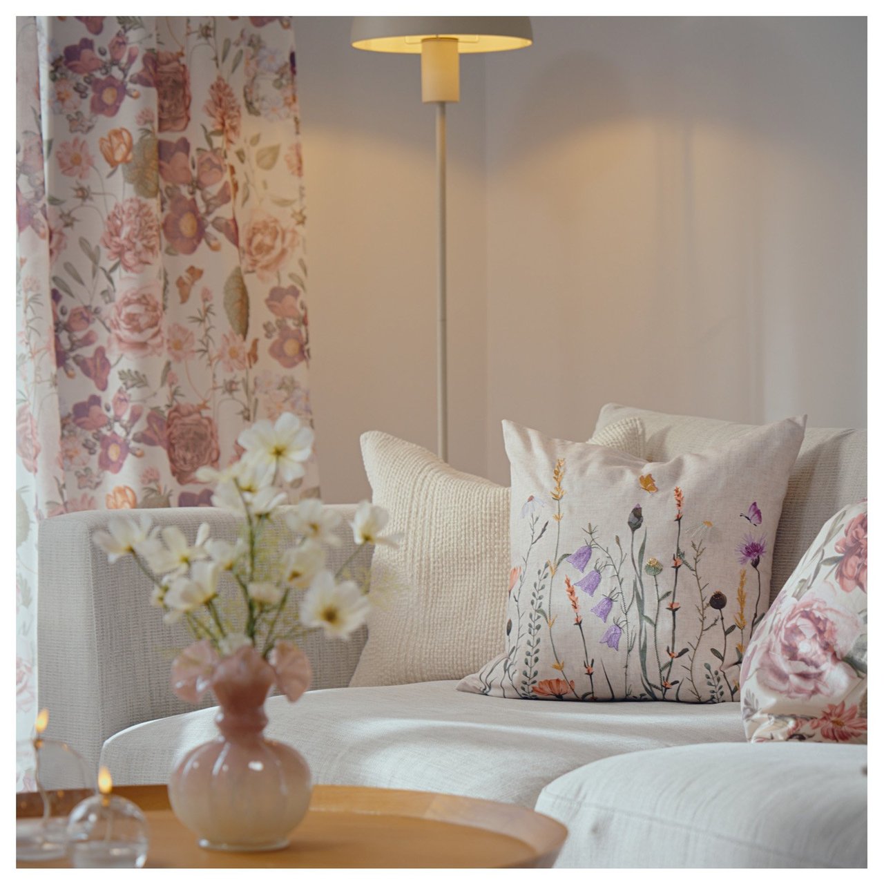 Olohuoneen nurkkaus, jossa on sohva, kukalliset verhot, lattiavalaisin ja maljakossa leikkokukkia sohvapöydällä.