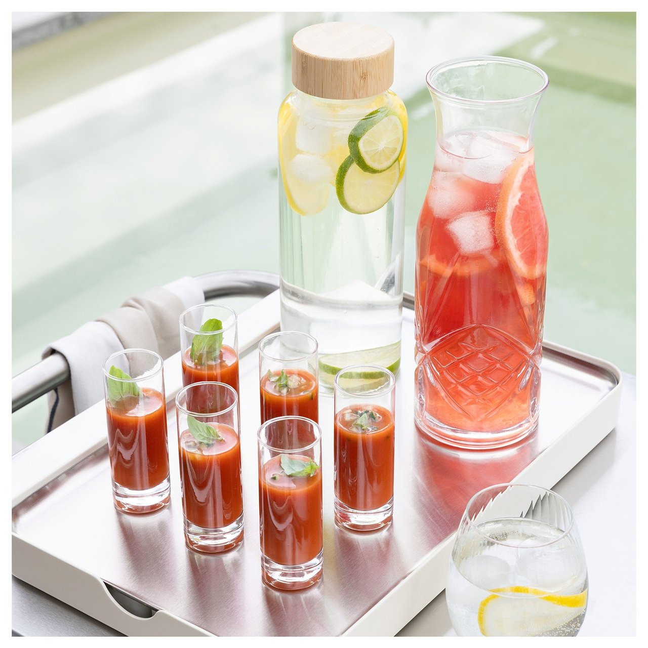 Seks små glass med gazpacho og to karafler står på et brett. Brettet står på en vogn. Bak vognen skimter man et svømmebasseng.
