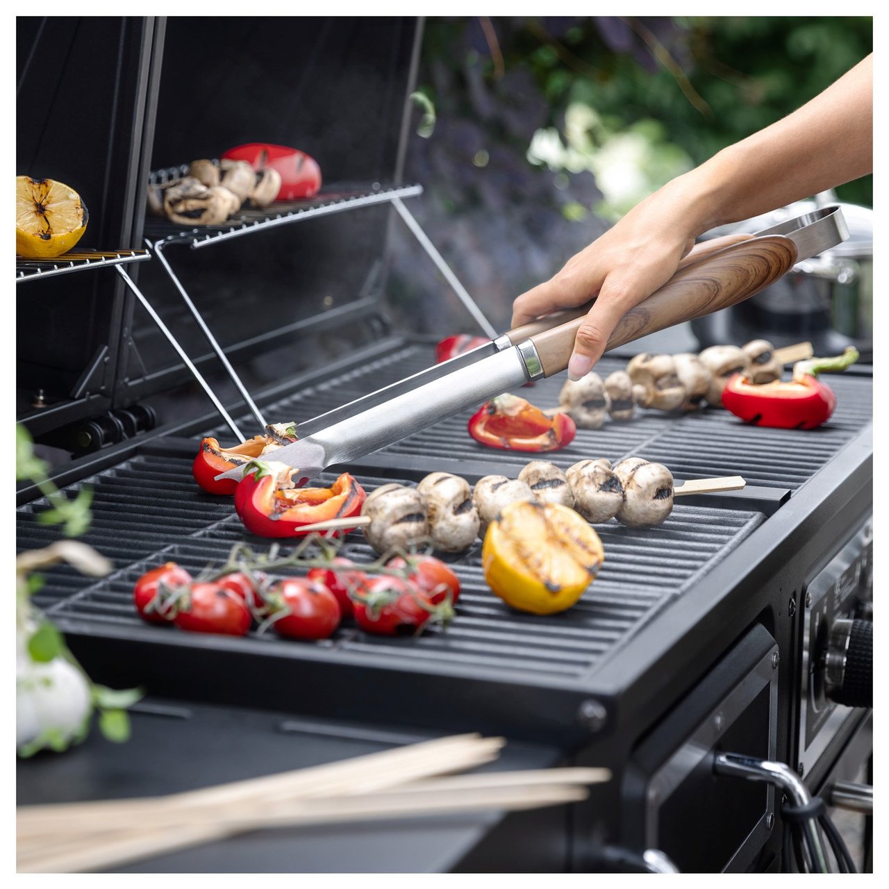 Grill mit Gemüse wie Paprika, Pilzen, Tomaten und Zitronen in Nahaufnahme. Eine Hand dreht mit einer Grillzange das Grillgut um.