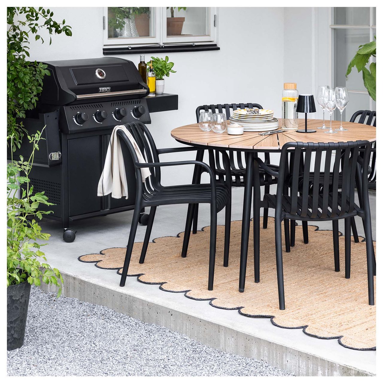 Uteplass med spisebord i lyst tre og fire svarte stoler på et beige teppe. Ved siden av står det en svart grill og en svart parasoll.