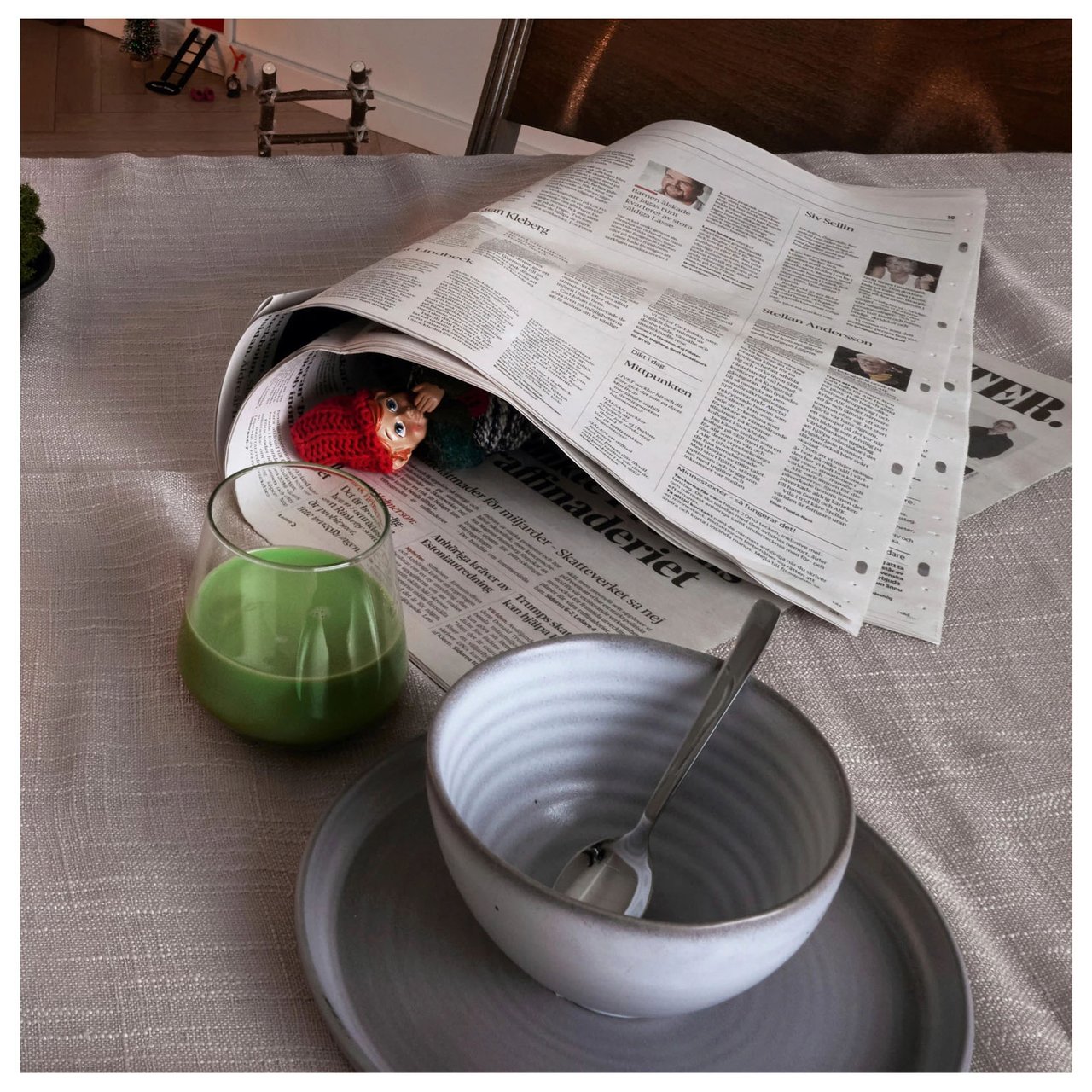 Ein kleiner schelmischer Wichtel versteckt sich in einer zusammengerollten Zeitung auf einem Frühstückstisch.