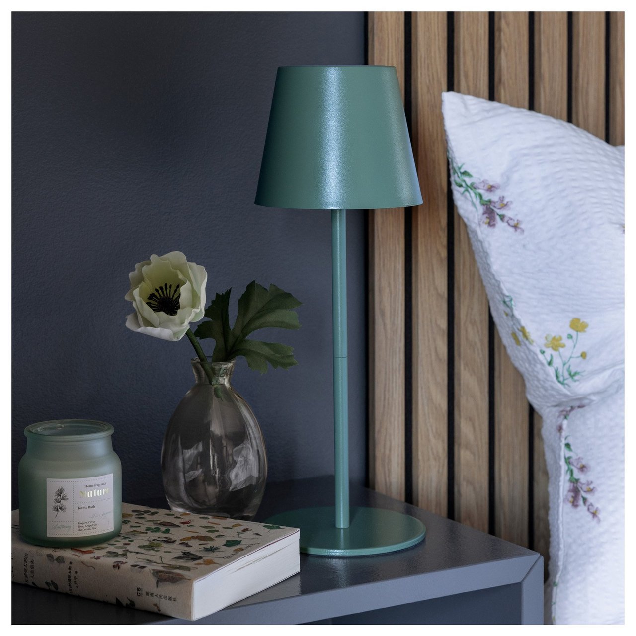 Nachttisch mit Lampe, Blumenvase, Duftkerzen und Buch neben einem Bett mit Akustikpaneelen als Kopfteil.