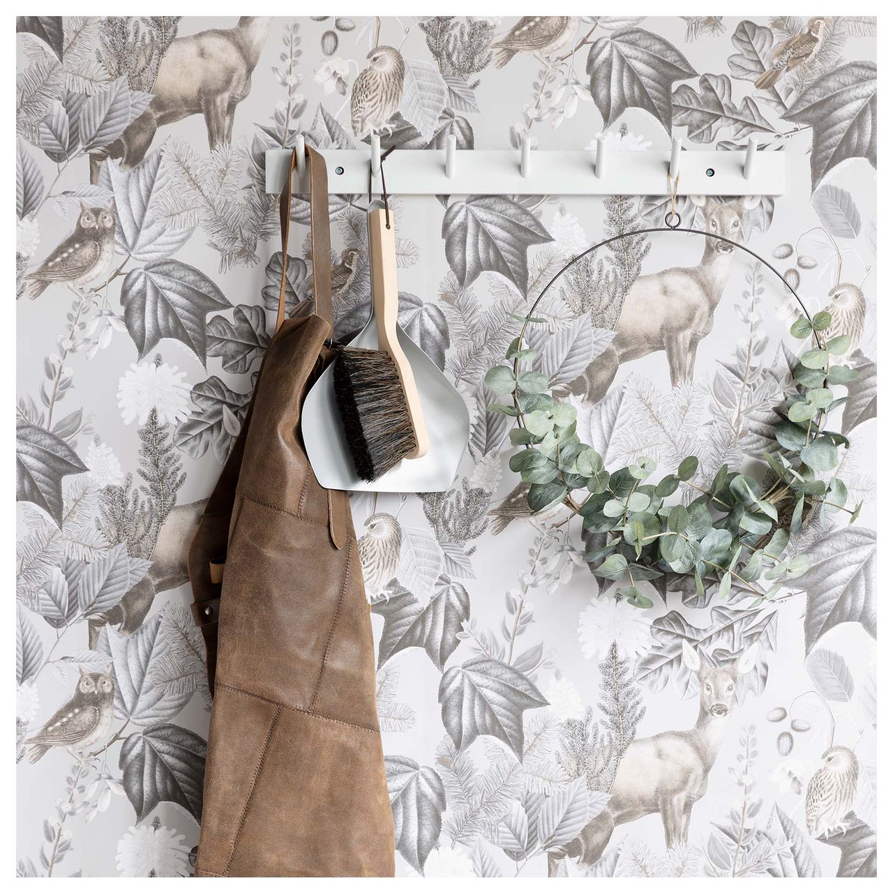 Vägg med kroklist där förkläde, borste och dekorativ krans hänger mot en naturinspirerad tapet.