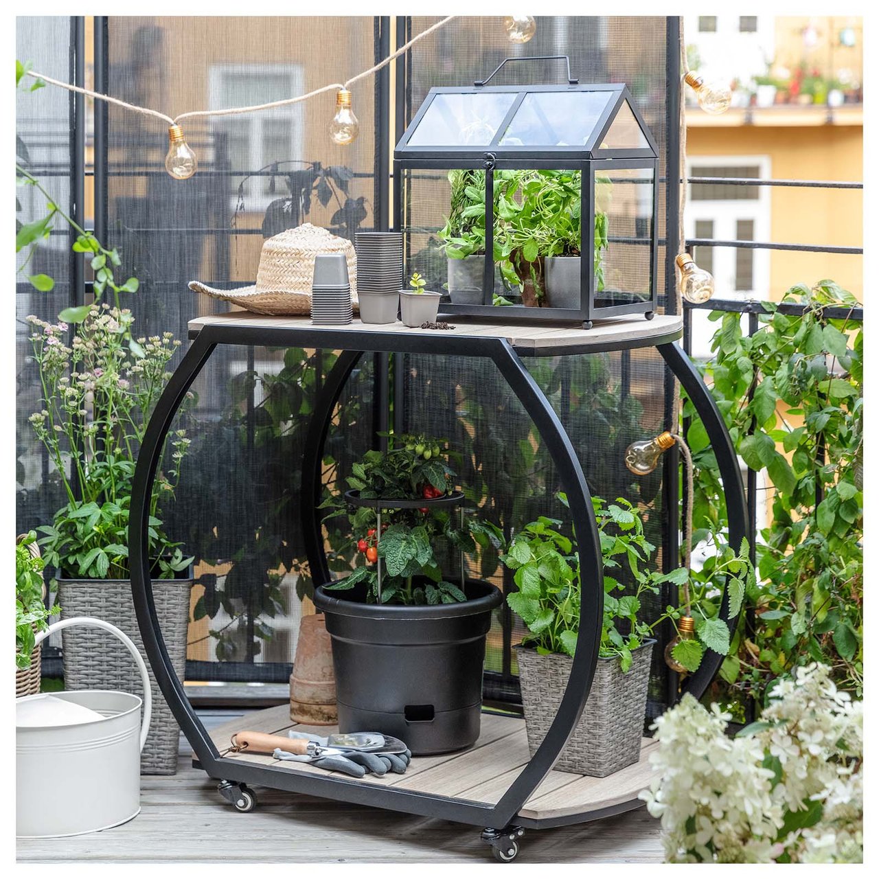Vogn med hjul som inneholder krukker, dyrkingstilbehør og et minidrivhus på en frodig balkong.