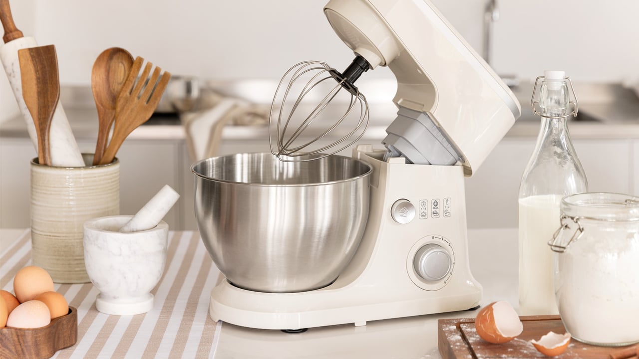 Köksassistent i beige med visp, omgiven av ingredienser och köksredskap i ett ljust kök.