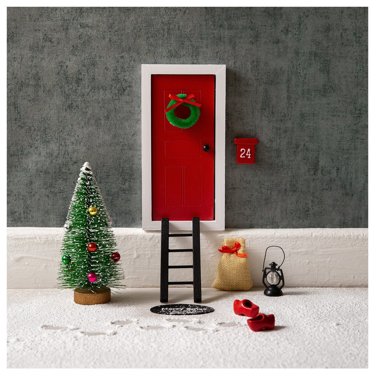 Rote Miniatur-Wichteltür mit Weihnachtskranz, Tannenbaum, Sack und roten Pantoffeln in winterlicher Weihnachtsszene.