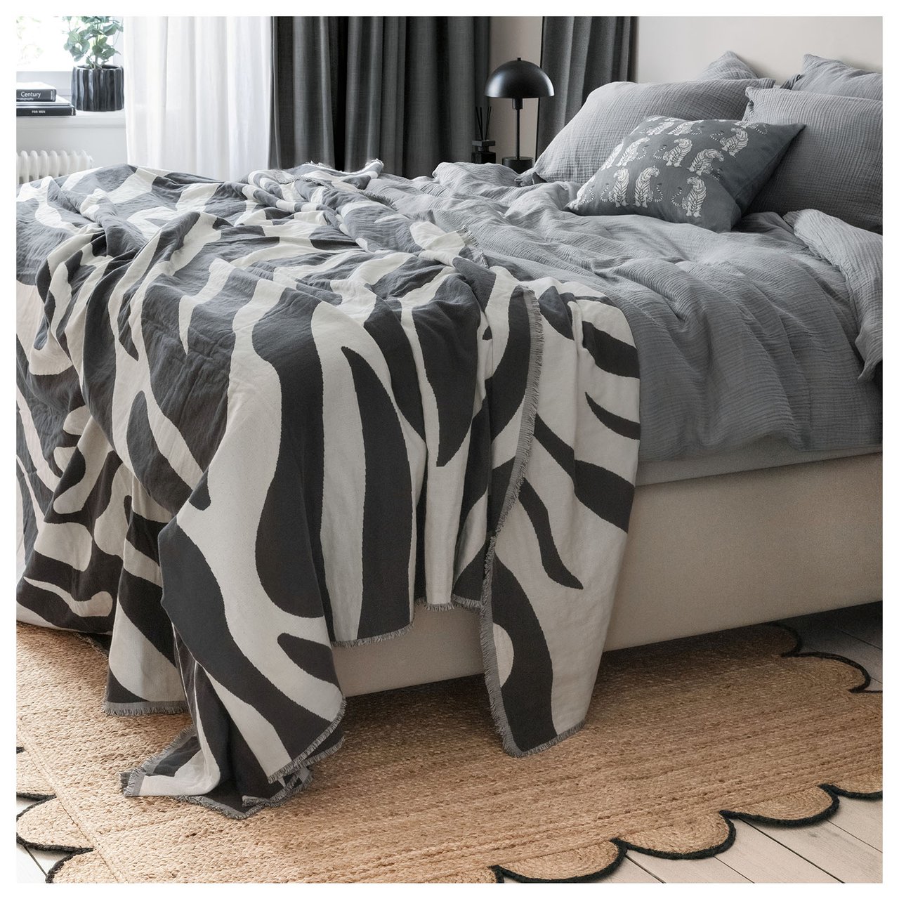Soverom med grått sengetøy, sebrastripet teppe og beige matte med svart kant.