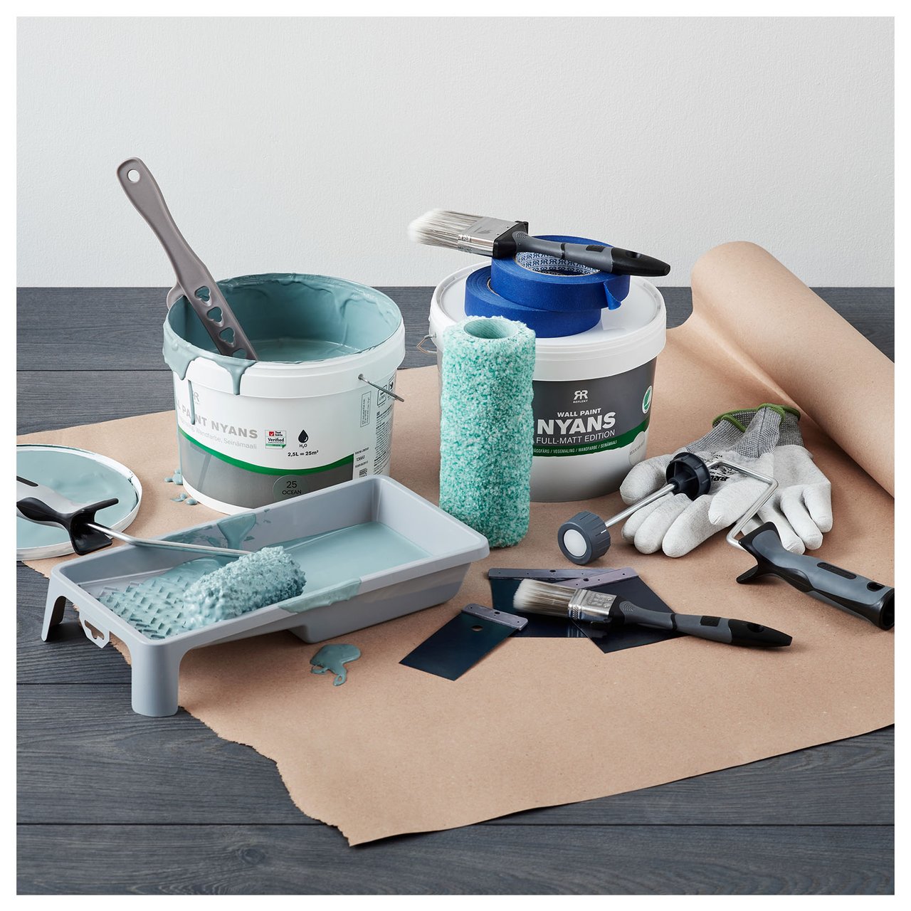 Maalausprojekti, jossa näkyy maalipurkkeja, teloja, siveltimiä, käsineitä ja tarvikkeita lattialla suojapaperin päällä.