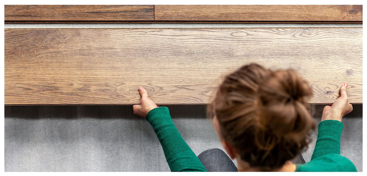 Närbild på kvinna med brunt hår och grön tröja som lägger laminatgolv i trä.