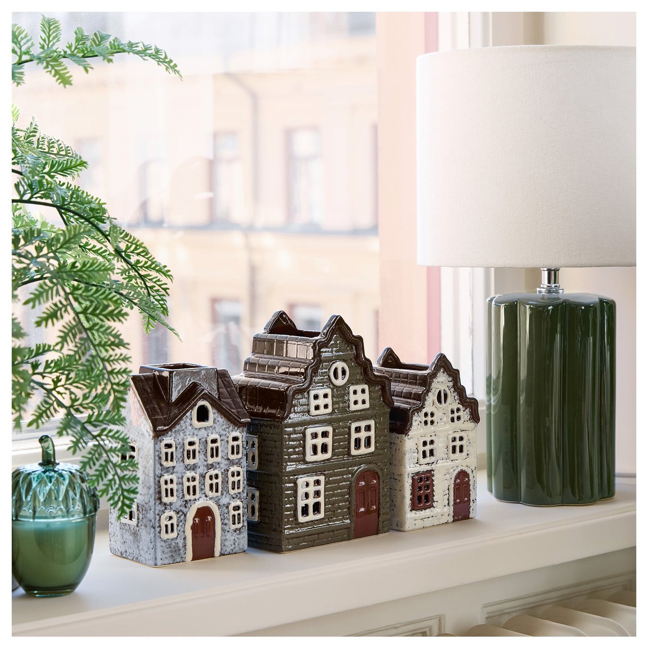Fönster med en bordslampa med grön keramikfot och vit tygskärm bredvid små ljuslyktor formade som hus.