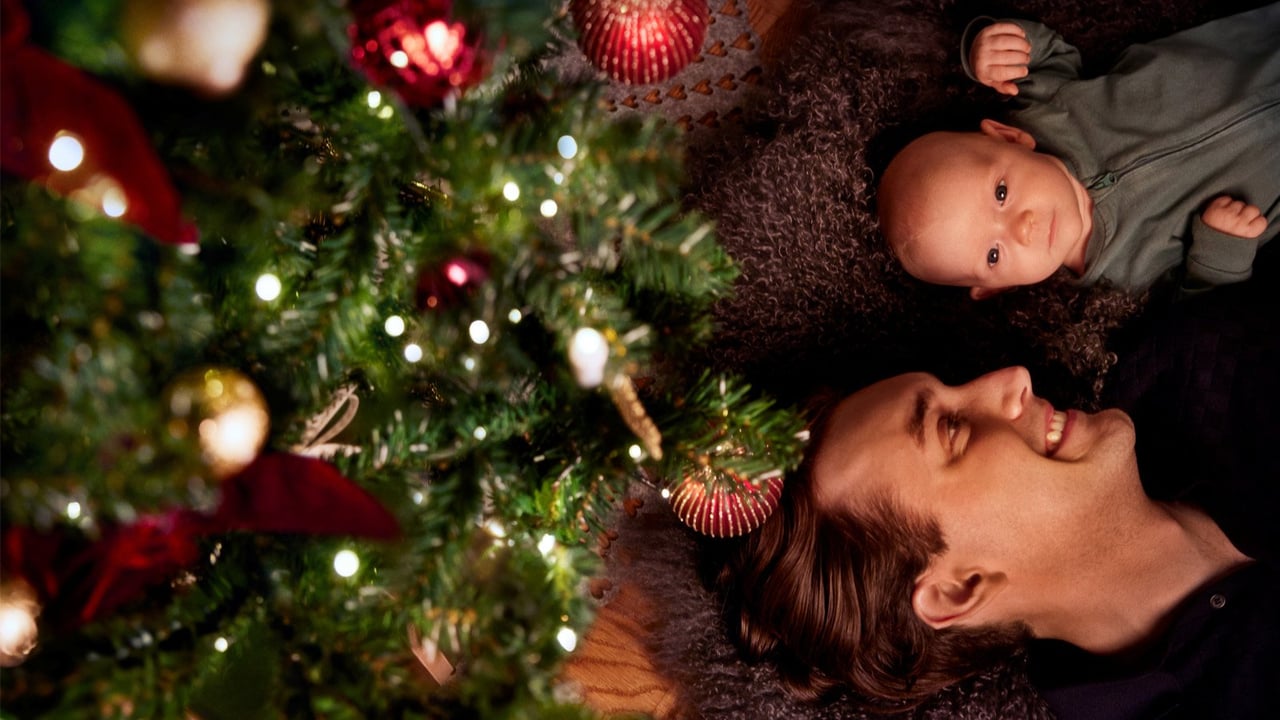 En man och en bebis ligger på golvet intill en pyntad julgran. Mannen tittar kärleksfullt på bebisen.