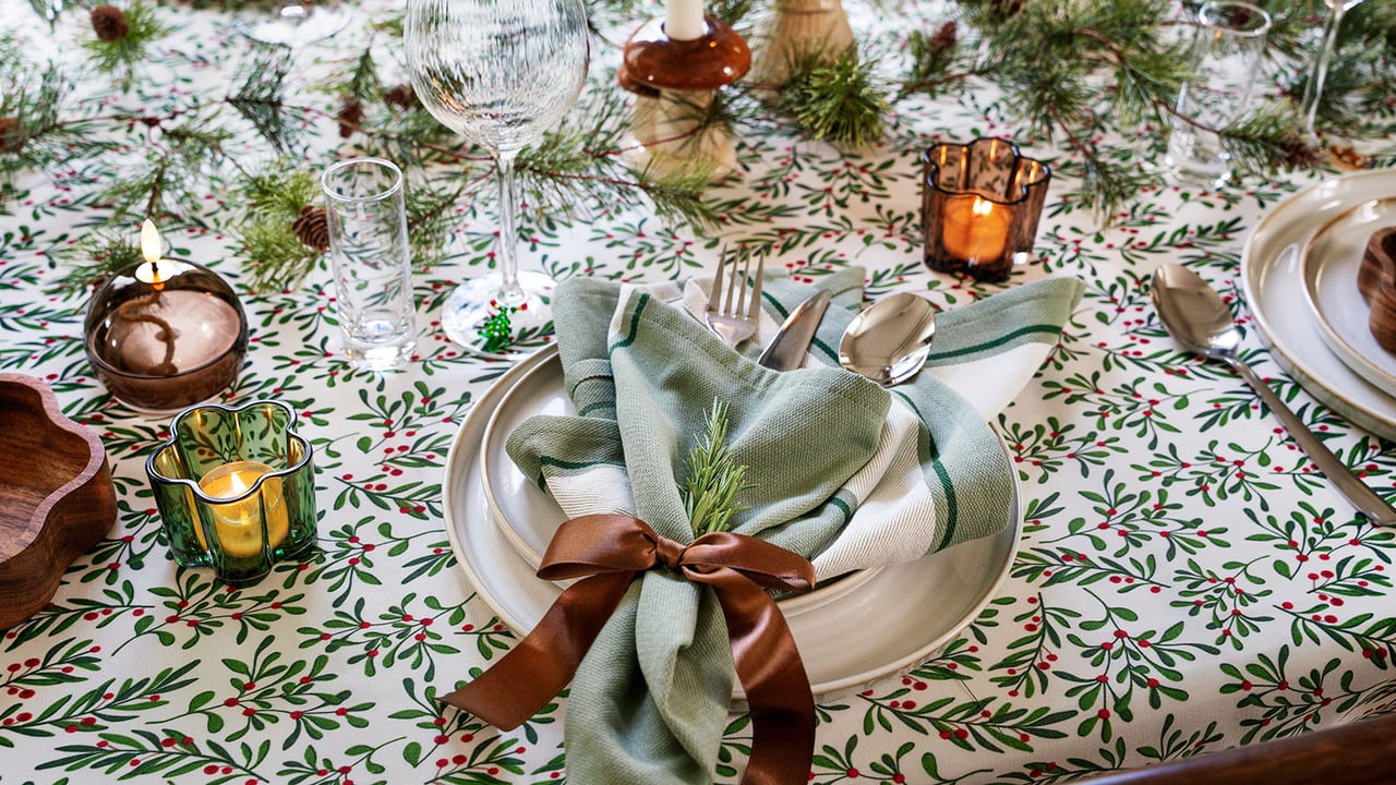 Weihnachtstisch mit grün-rotem gemustertem Tuch, Tellern, Leinenserviette mit Schleife und Kerzen.