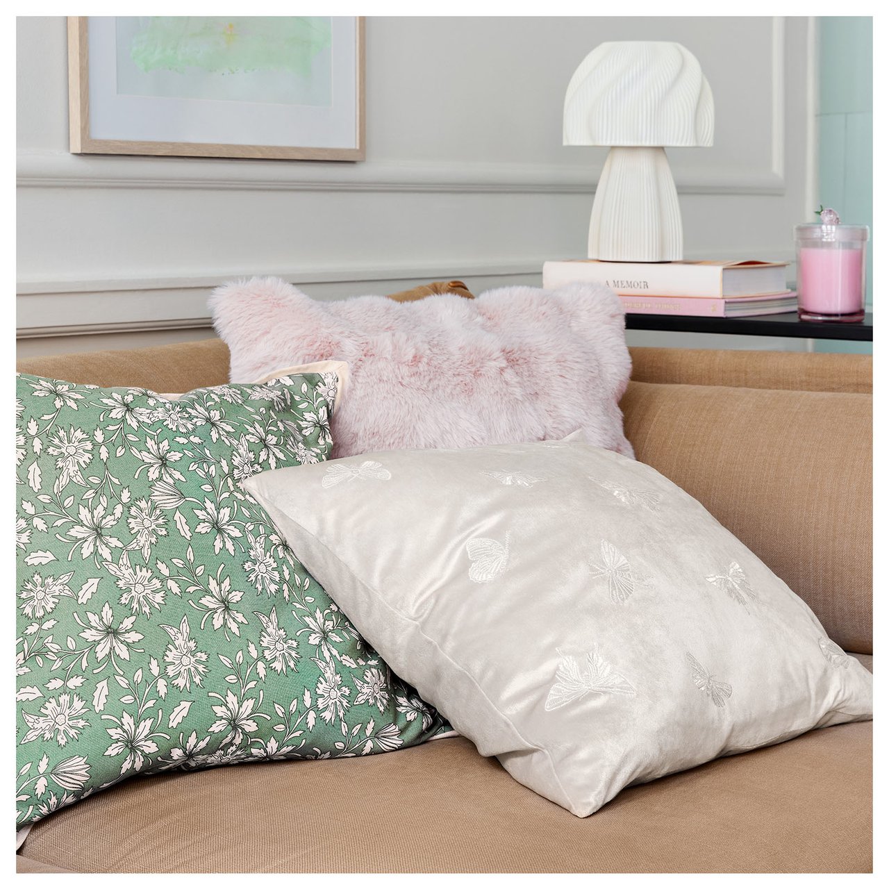 Tre pastellfargede puter ligger i en sofakrok. Bak står det en hvit lampe på et sidebord.