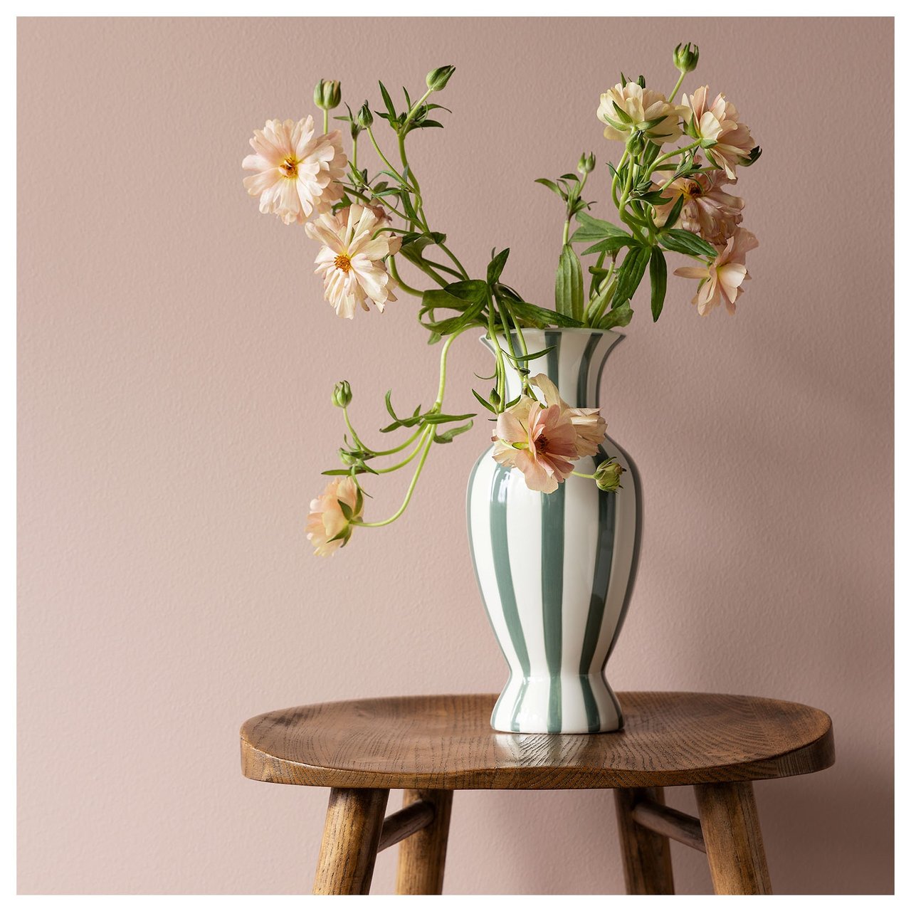 Pieni puinen pöytä, jolla on raidallinen maljakko ja kukka-asetelma hennon vaaleanpunaisen sävyisen seinän edessä.