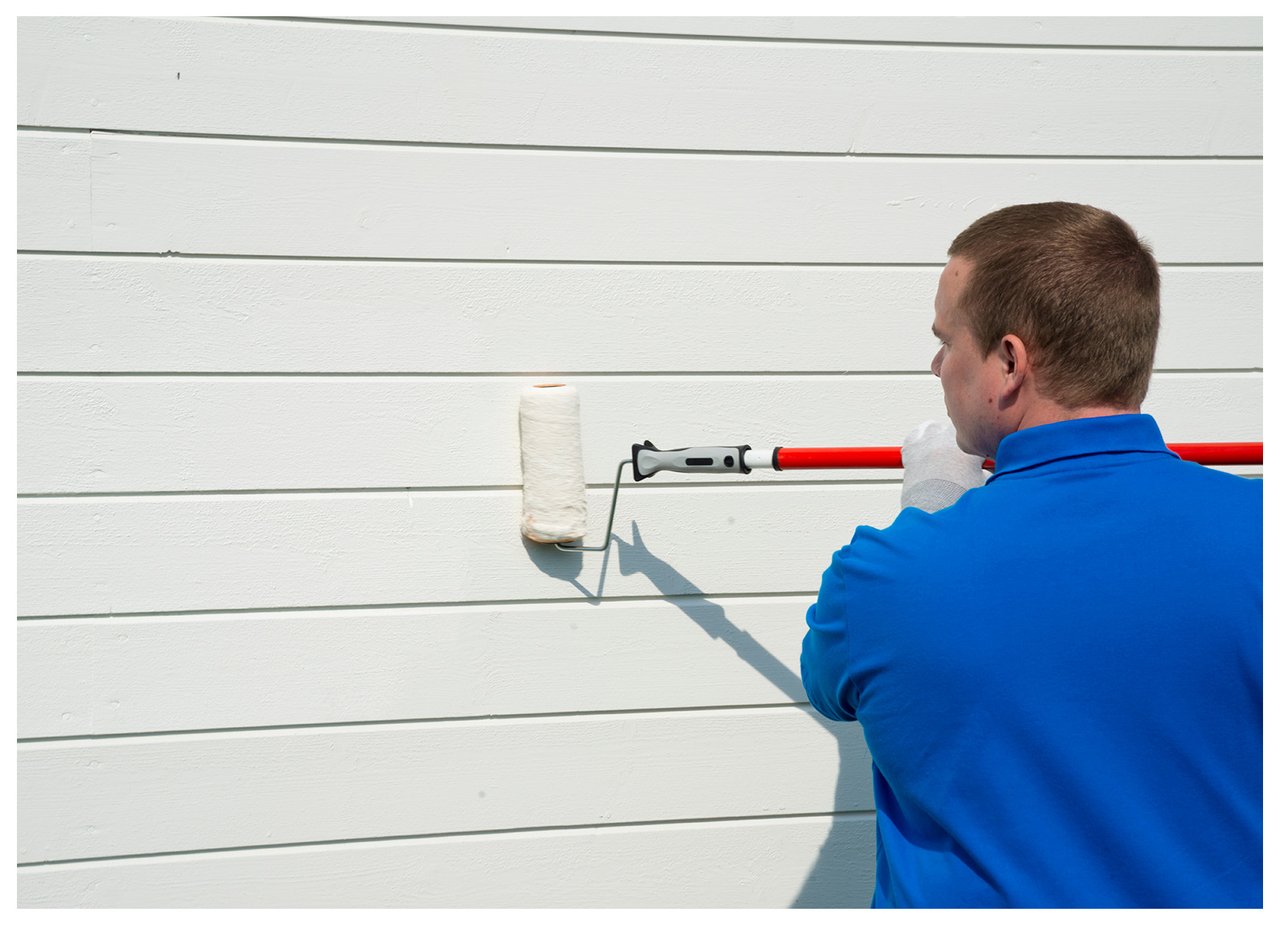 Man med blå t-shirt målar en vit husfasad med roller som har ett förlängningsskaft.