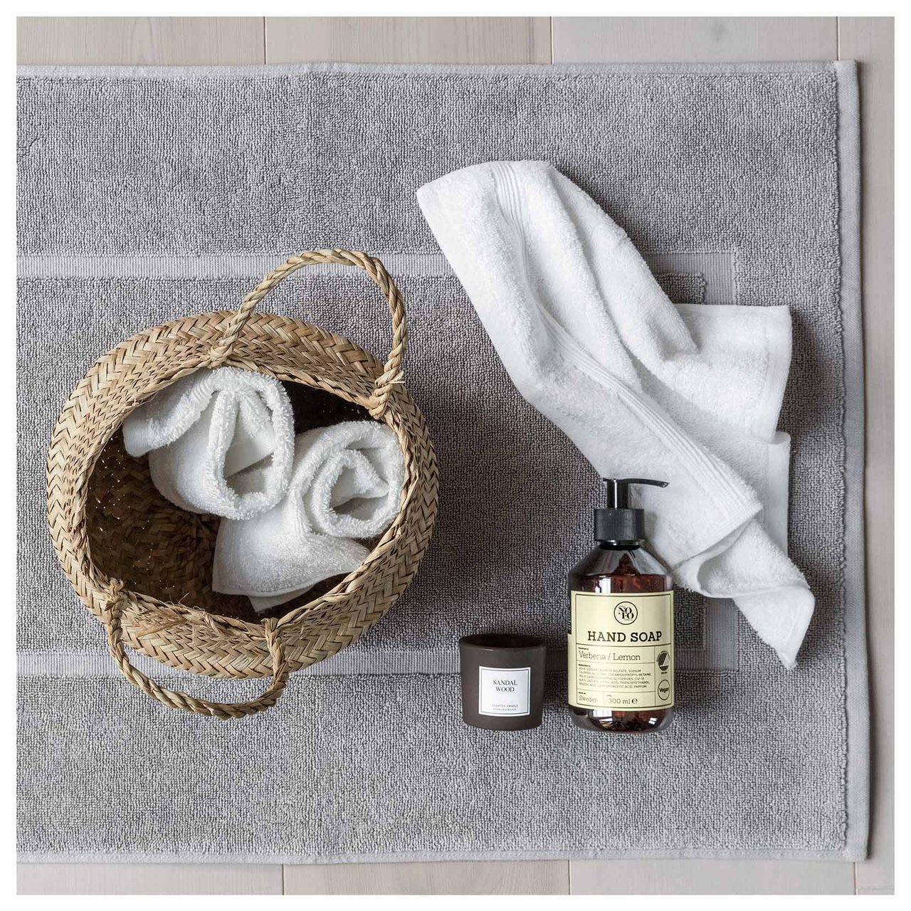 Grå baderomsmatte med flytende såpe, et duftlys og en kurv fylt med rullede håndklær.
