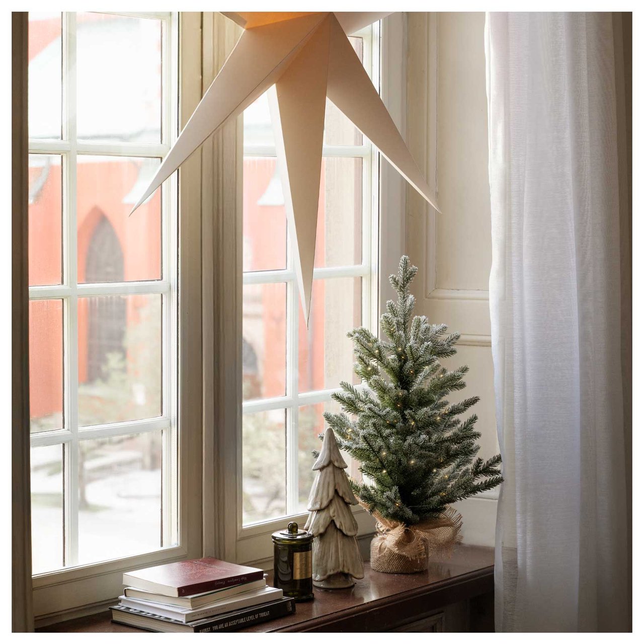 Valkoinen adventtitähti roikkuu ikkunassa. Ikkunalaudalla on pieniä koristekuusia.