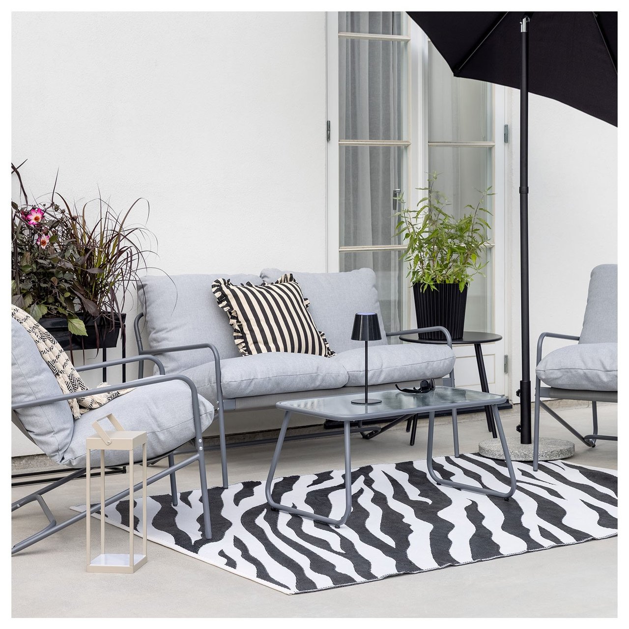 Kompaktes Lounge-Set aus schwarzem Stahl, bestehend aus einem Sofa, zwei Sesseln und einem Tisch. Die Sitzgruppe steht unter einem Sonnenschirm auf einer Terrasse aus Steinplatten.