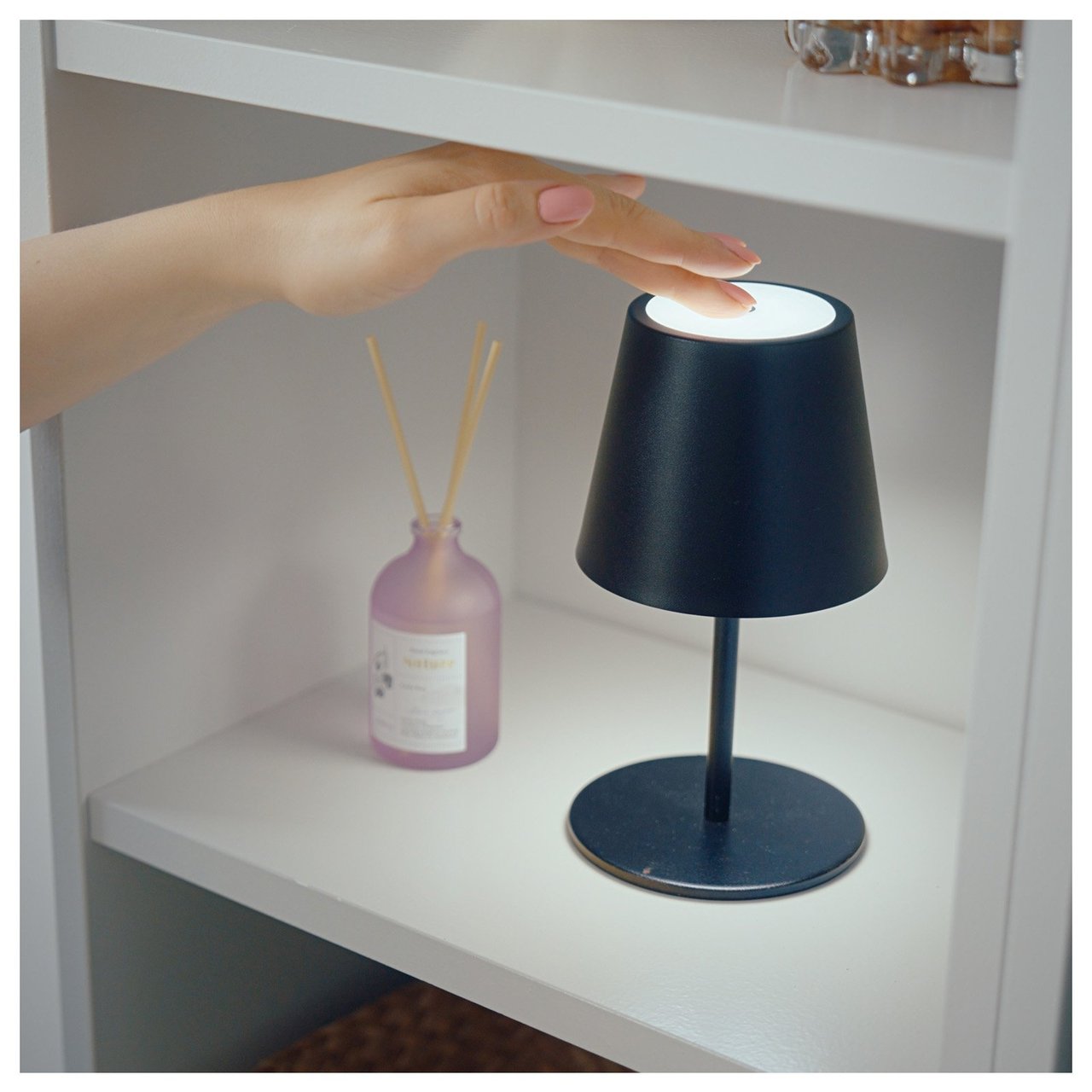 Käsi sytyttää pöytävalaisimen, joka on hyllyllä tuoksutikkuja sisältävän pullon vieressä.
