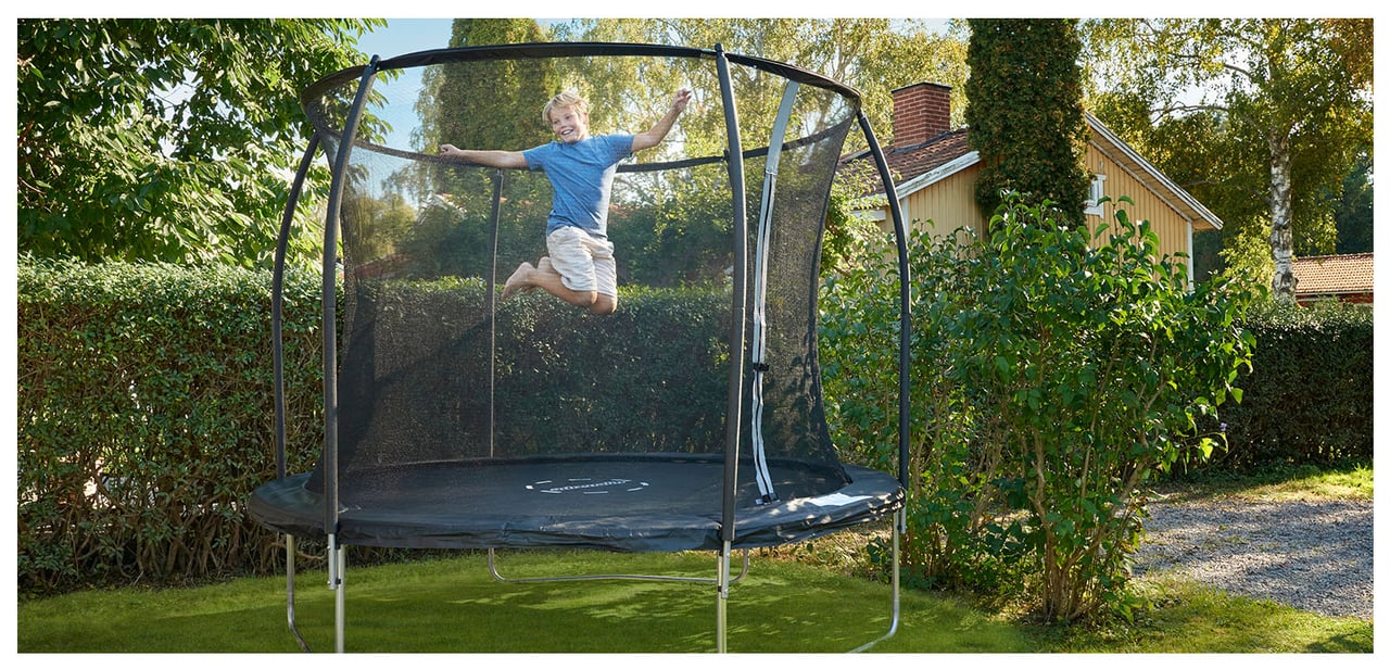 Gutt i blå t-skjorte og shorts hopper på en trampoline i en frodig hage.