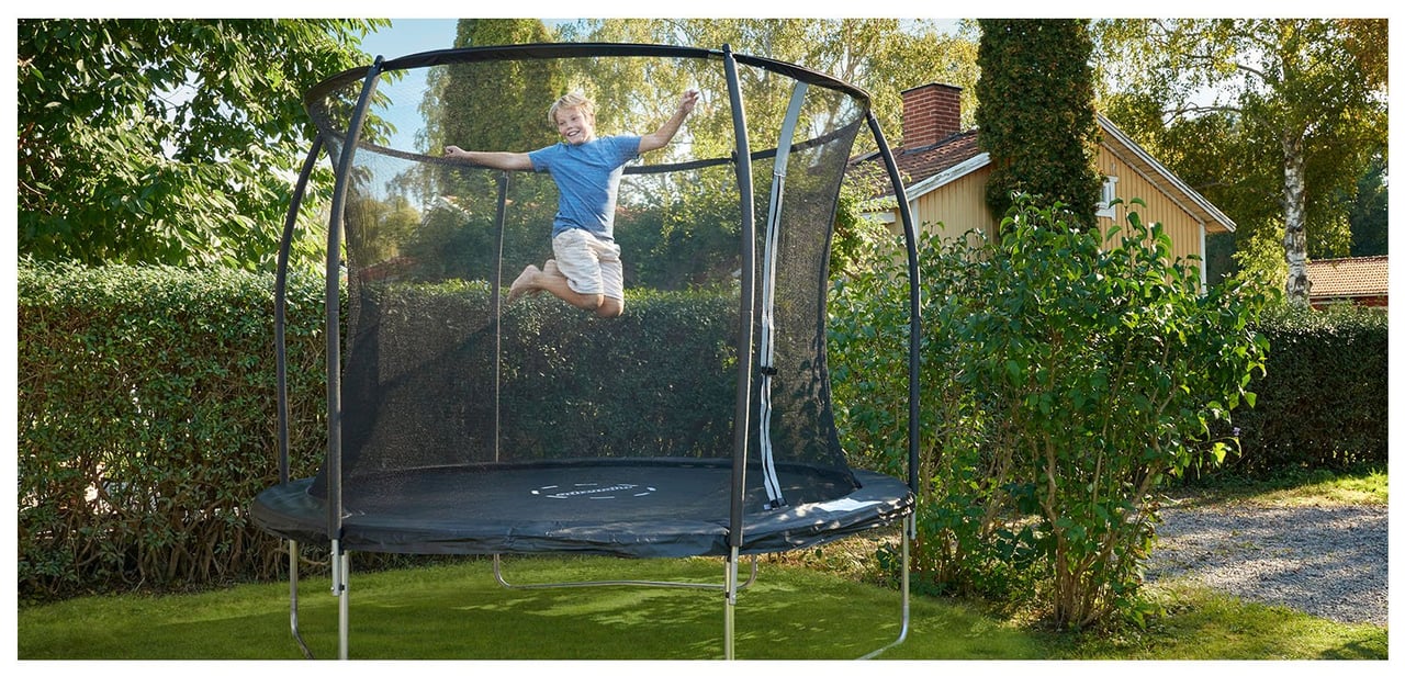 Gutt i blå t-skjorte og shorts hopper på en trampoline i en frodig hage.