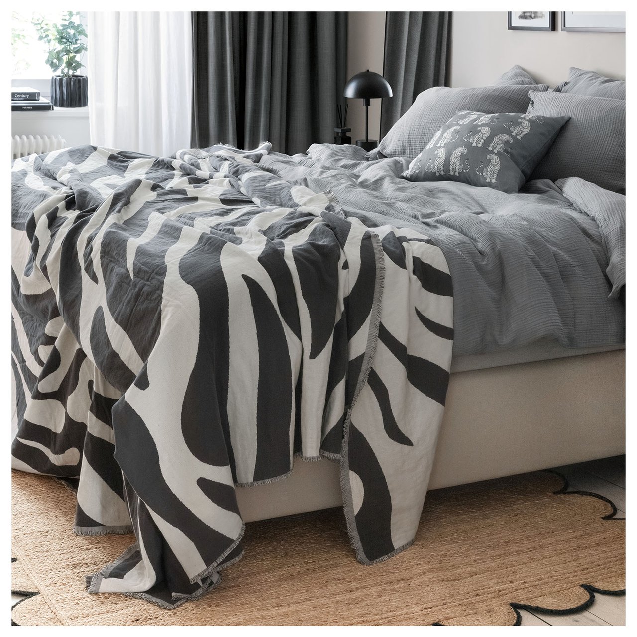 En dobbeltseng er redd opp med grå dynetrekk. Under sengen ligger det et juteteppe med svarte detaljer.