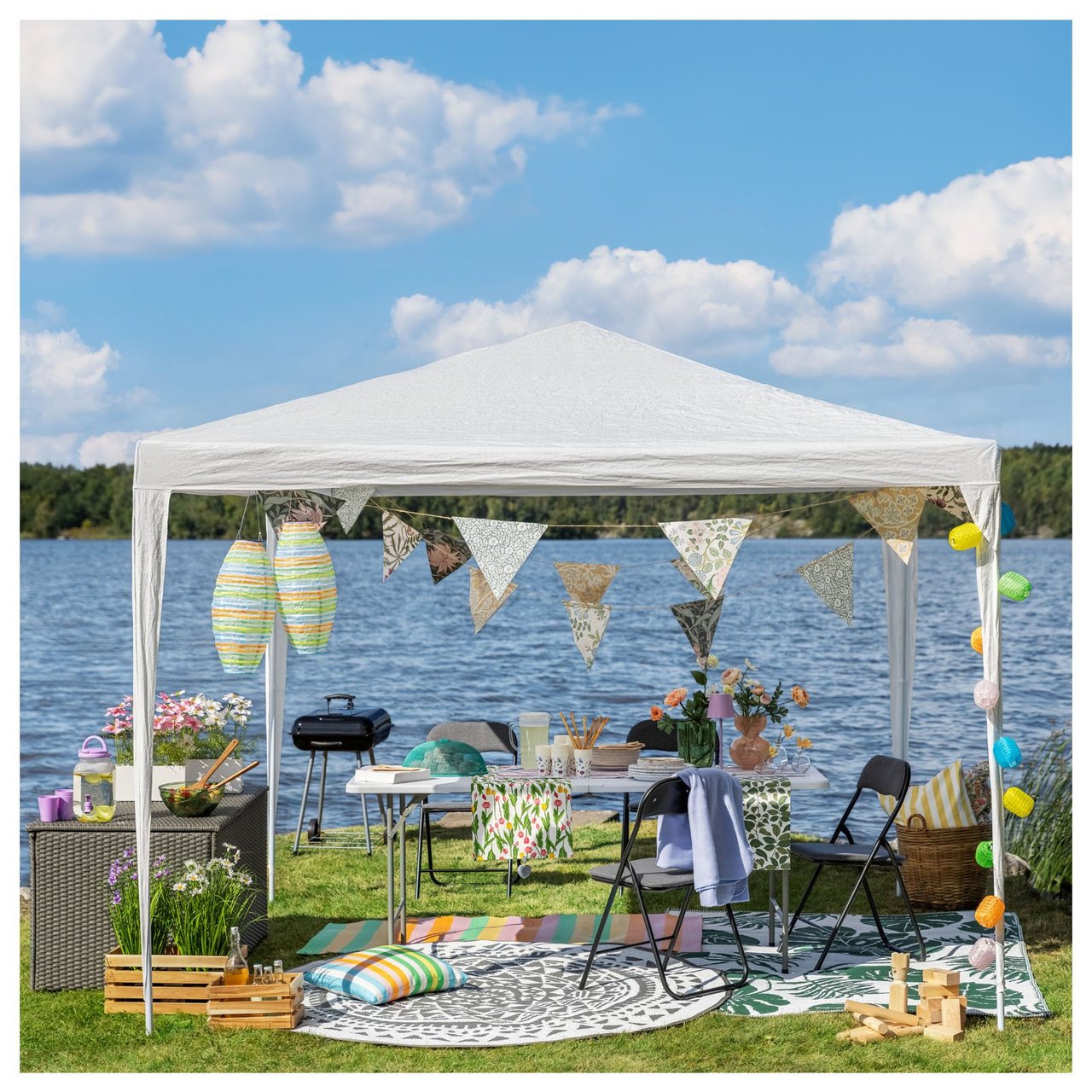 Ein stimmungsvolles Sommerfest am Wasser mit einem Partyzelt, das mit Wimpeln, Reislampen und Lichterketten dekoriert ist. Hier steht ein gedeckter Tisch mit Blumen und sommerlichen Details, umgeben von Stühlen, Kissen und Outdoor-Teppichen.