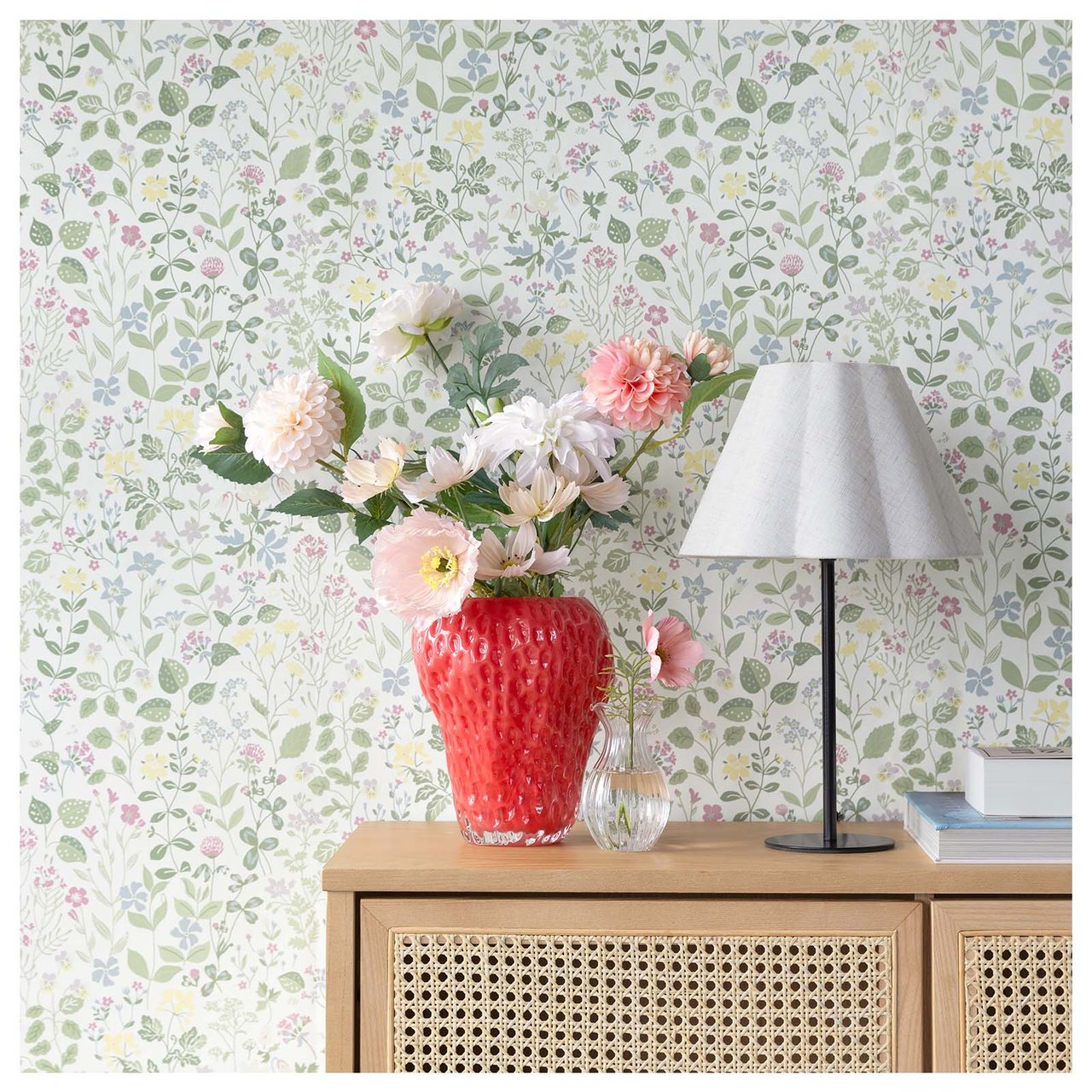 Småblommig tapet bakom ett skåp. På skåpet står röd vas med blommor och en svartvit lampa.