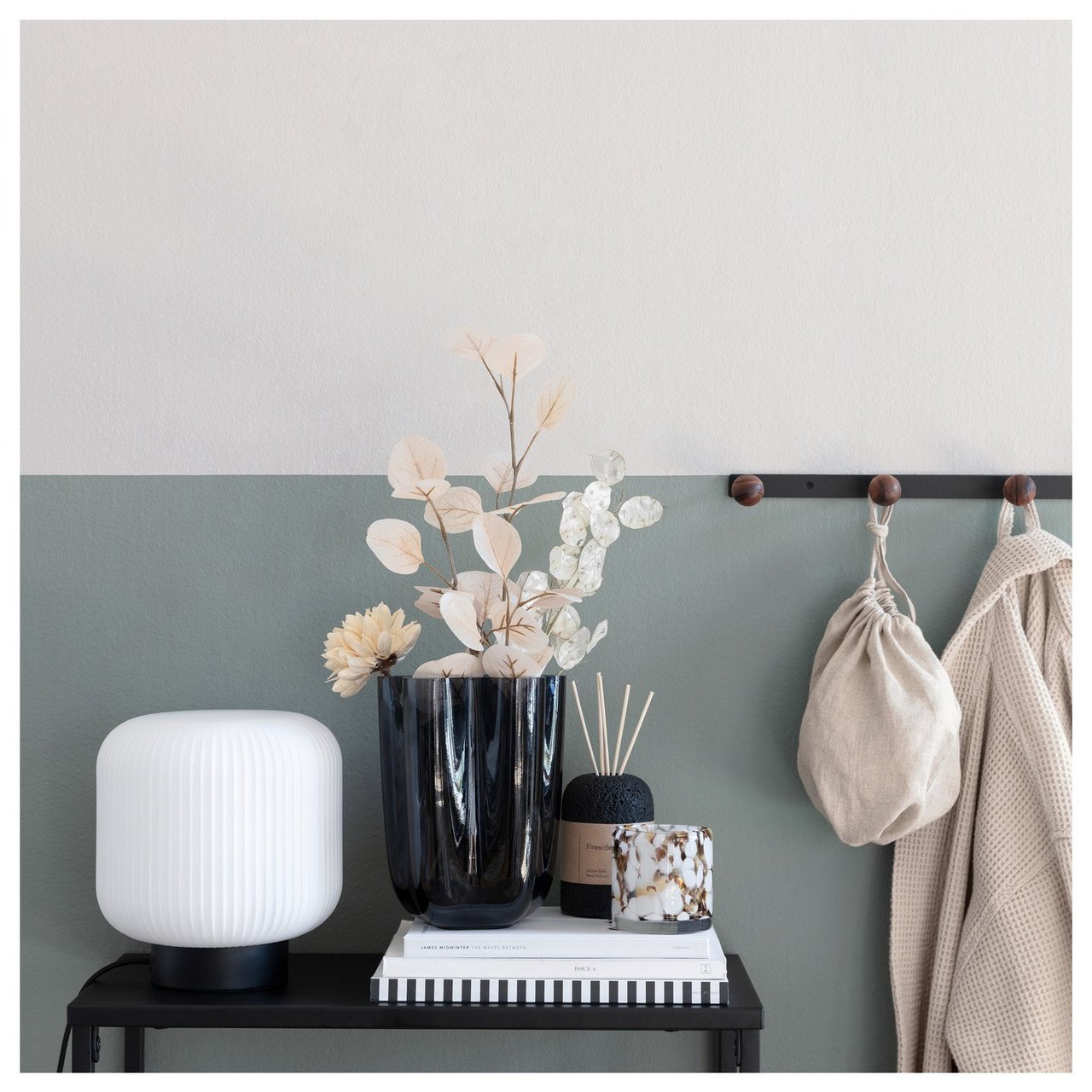 Lun gang med bord, dekorativ belysning, vase med tørkede blomster og knaggrekke på veggen.