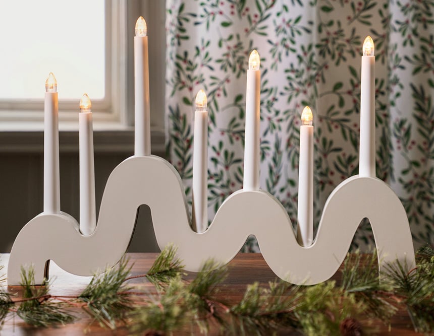 Ein elektrischer Adventsleuchter mit sieben Kerzen steht auf einem Tisch vor einem Fenster mit Weihnachtsdekoration.