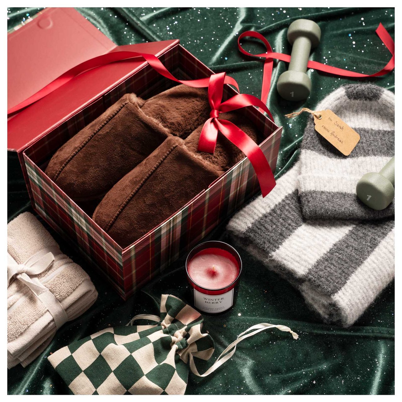 Ein Paar braune Hausschuhe in einer Geschenkbox. Daneben stehen eine Duftkerze und kleine Handtücher.