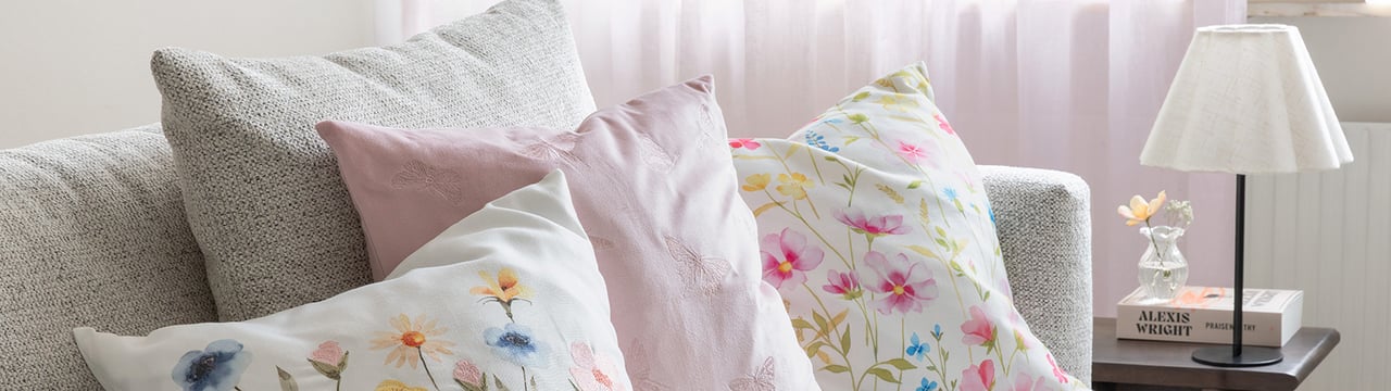 Lys sofa med dekorative puter i pastellfarger og blomstermønster ved siden av et lite bord med lampe.