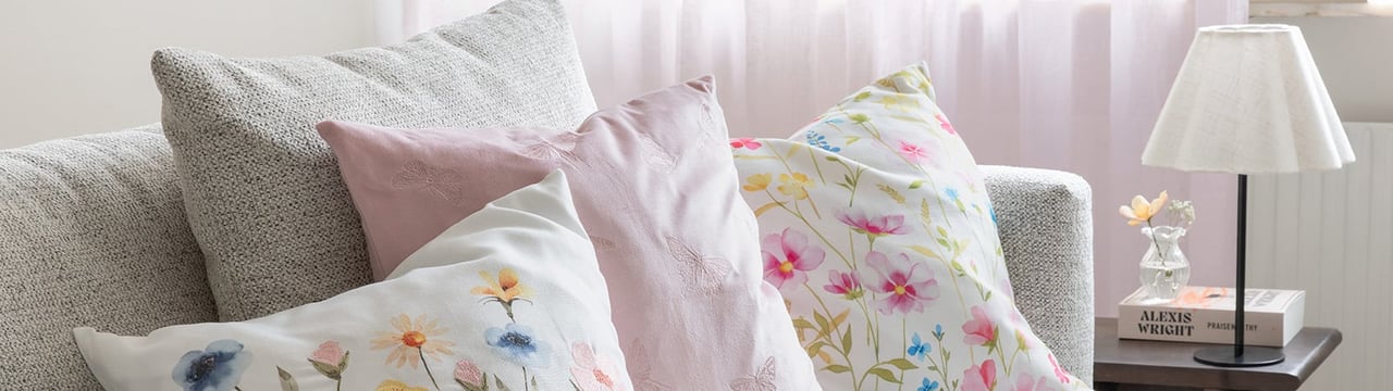 Lys sofa med dekorative puter i pastellfarger og blomstermønster ved siden av et lite bord med lampe.