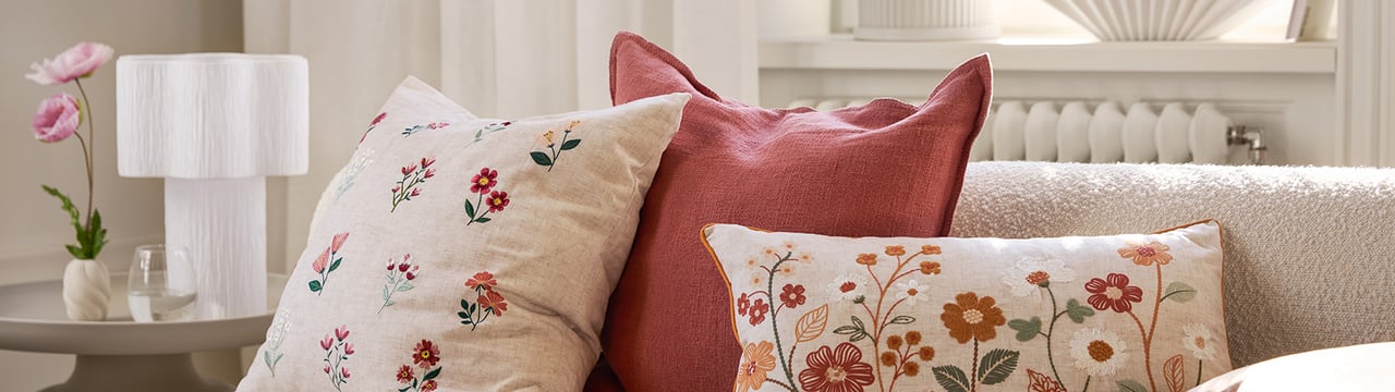 Lys sofa med blomstrete pynteputer i varme toner, hvit bordlampe og grønn plante i en solfylt stue.