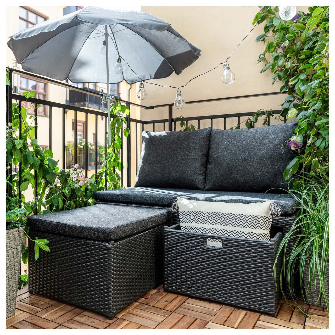 Hjørnedel av balkong i bymiljø med loungesofa i svart kunstrotting og mørkegrå parasoll.