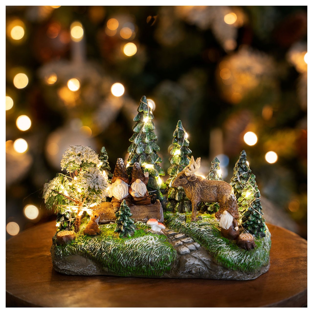 Kleines, beleuchtetes Weihnachtsdorf mit Bäumen, Figuren und Elchen auf einem Tisch vor einem Weihnachtsbaum.