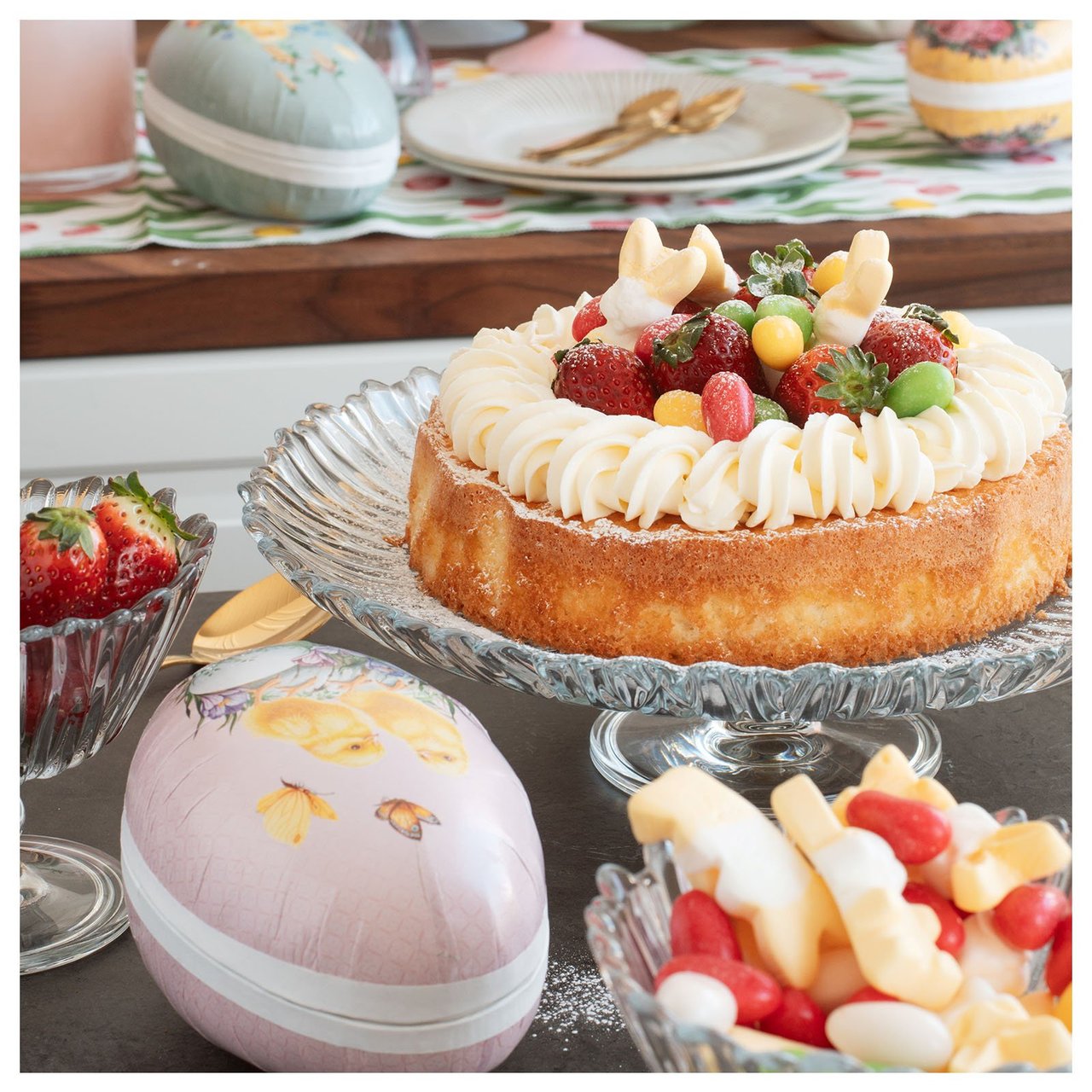 Gedeckter Ostertisch mit einem Kuchen auf einem Glasteller, umgeben von Kerzen, sowie Osterzweige in einer hellrosa Glasvase.