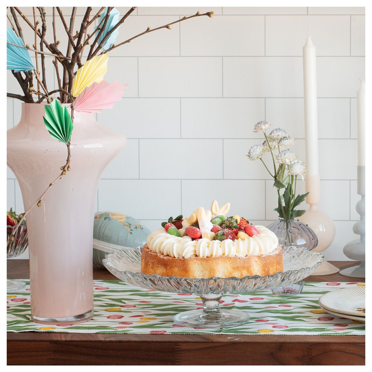 Gedeckter Ostertisch mit einem Kuchen auf einem Glasteller, umgeben von Kerzen, sowie Osterzweige in einer hellrosa Glasvase.