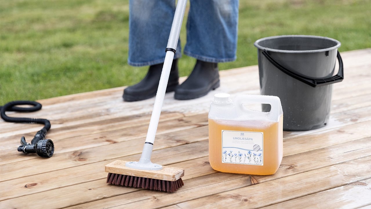 Eine Person reinigt eine Holzterrasse mit Leinölseife und einer Bürste. Auch ein Eimer und ein Gartenschlauch sind zu sehen.