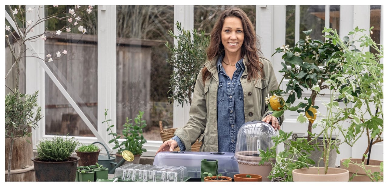 Sofia Björk står i drivhuset sitt foran en benk med krukker, drivhus, frø og planter.