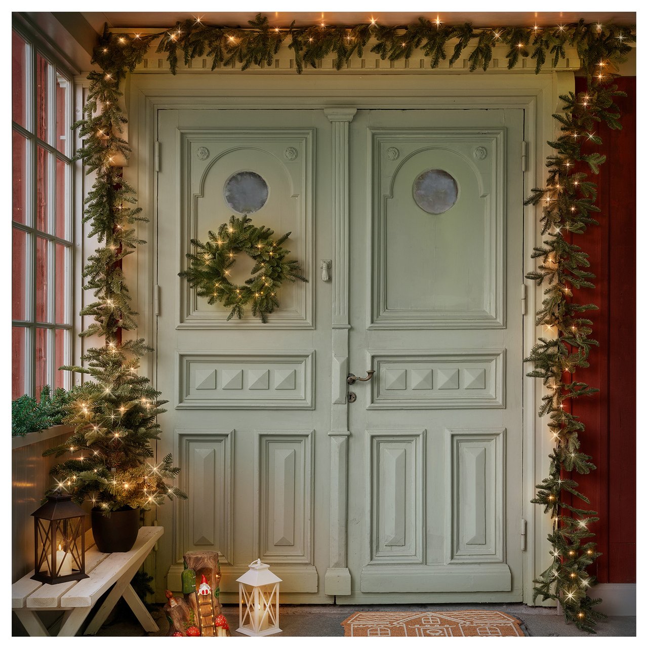 Juldekorerad ytterdörr med krans, belysning och pyntade granar på sidorna.