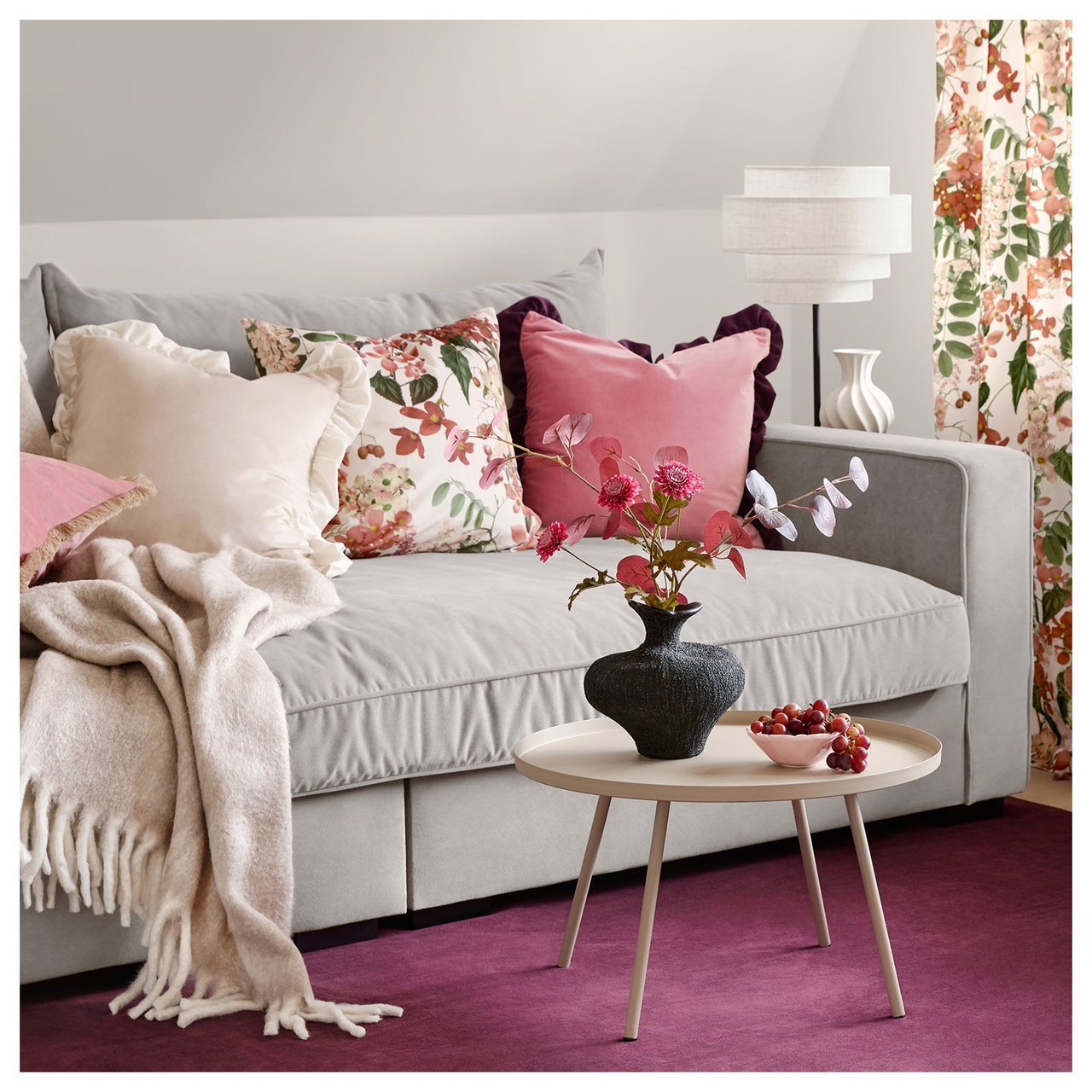 Wohnzimmer mit grauem Sofa, rosa Kissen, gemusterten Vorhängen und rosa Teppich.