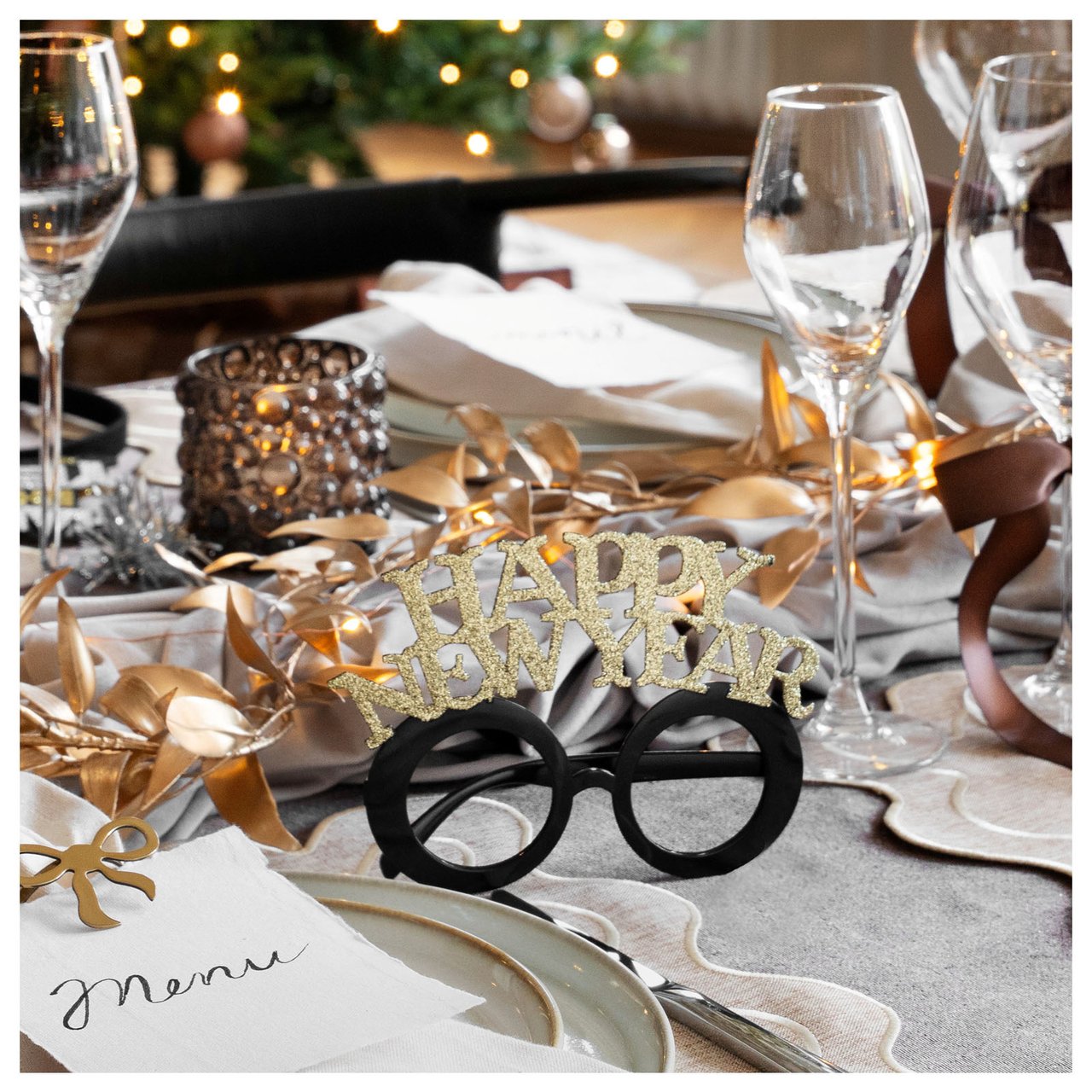 Nyårsdukning med glas, tallrikar, ljusslinga och festglasögon med guldfärgad text ”Happy New Year”.