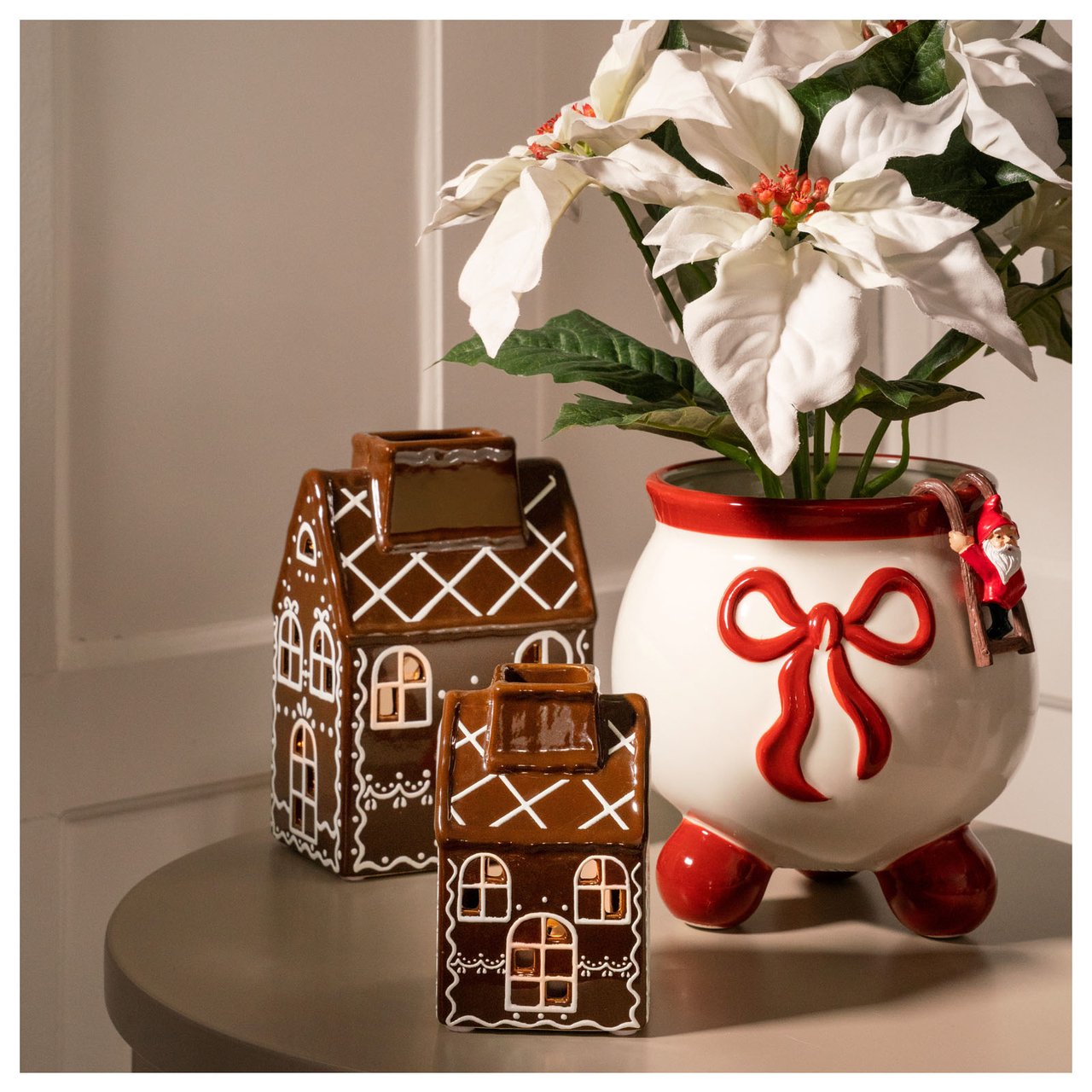 Ljuslyktor i form av pepparkakshus och en julblomma i kruka med röd rosett.