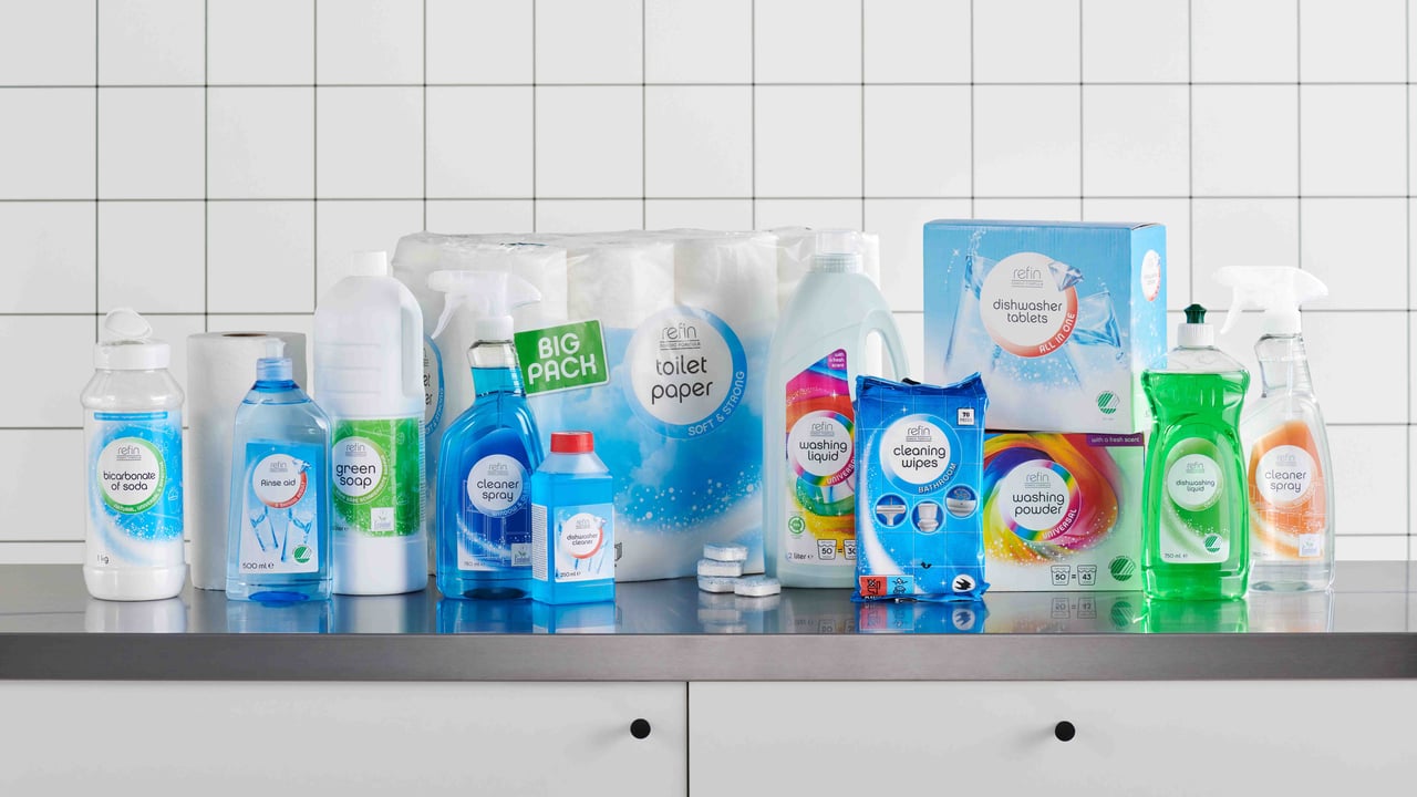 Reinigungsprodukte und Toilettenpapier, aufgestellt auf einem weißen Tisch vor einer gefliesten Wand.